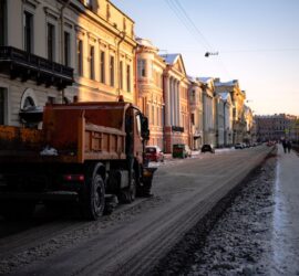 Orange snow-clearing truck on a snow-covered city street at sunset, pastel historic buildings lining the block to the left and a canal to the right
