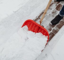 Person in jeans and black boot pushes a red snow shovel to clear snow from a brick sidewalk outdoors.