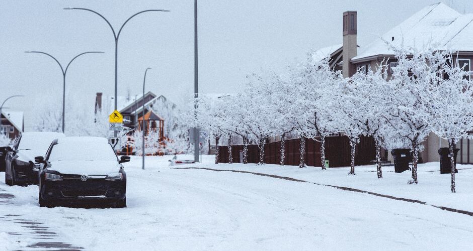 Snow-covered residential street with parked cars, snow-laden trees, and houses along the curb under a pale winter sky, including a yellow pedestrian crossing sign nearby.