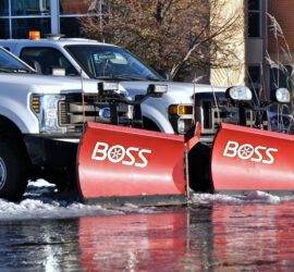 Two white pickup trucks with mounted red BOSS snow plows parked on a wet, snowy lot.